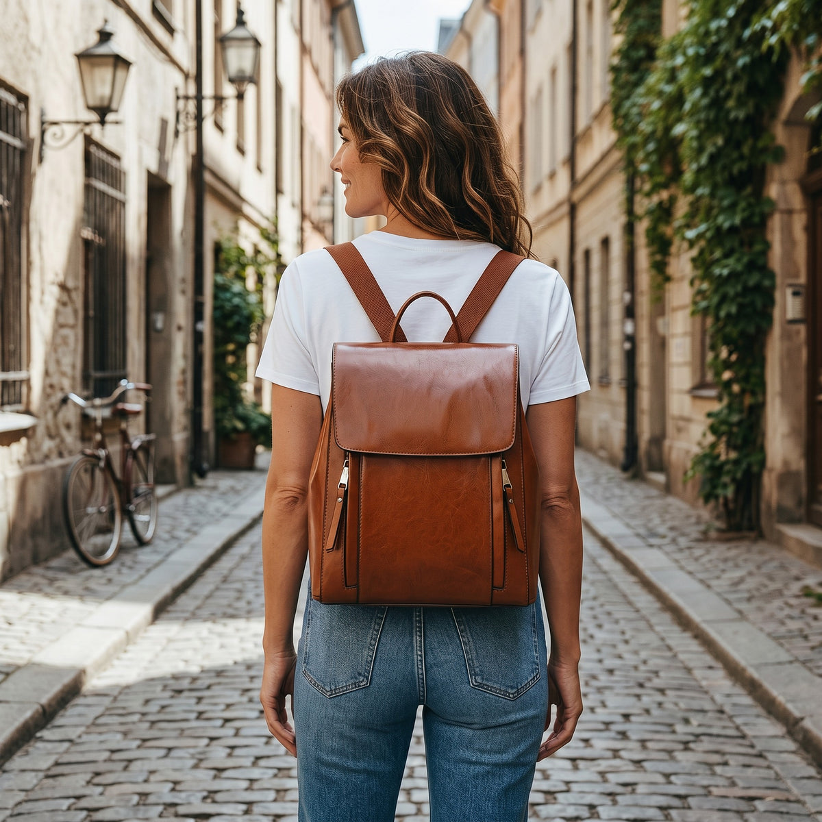 Femme portant un sac à dos bandoulière Suali en cuir marron Le Voyage Prestige dans une rue pavée européenne.